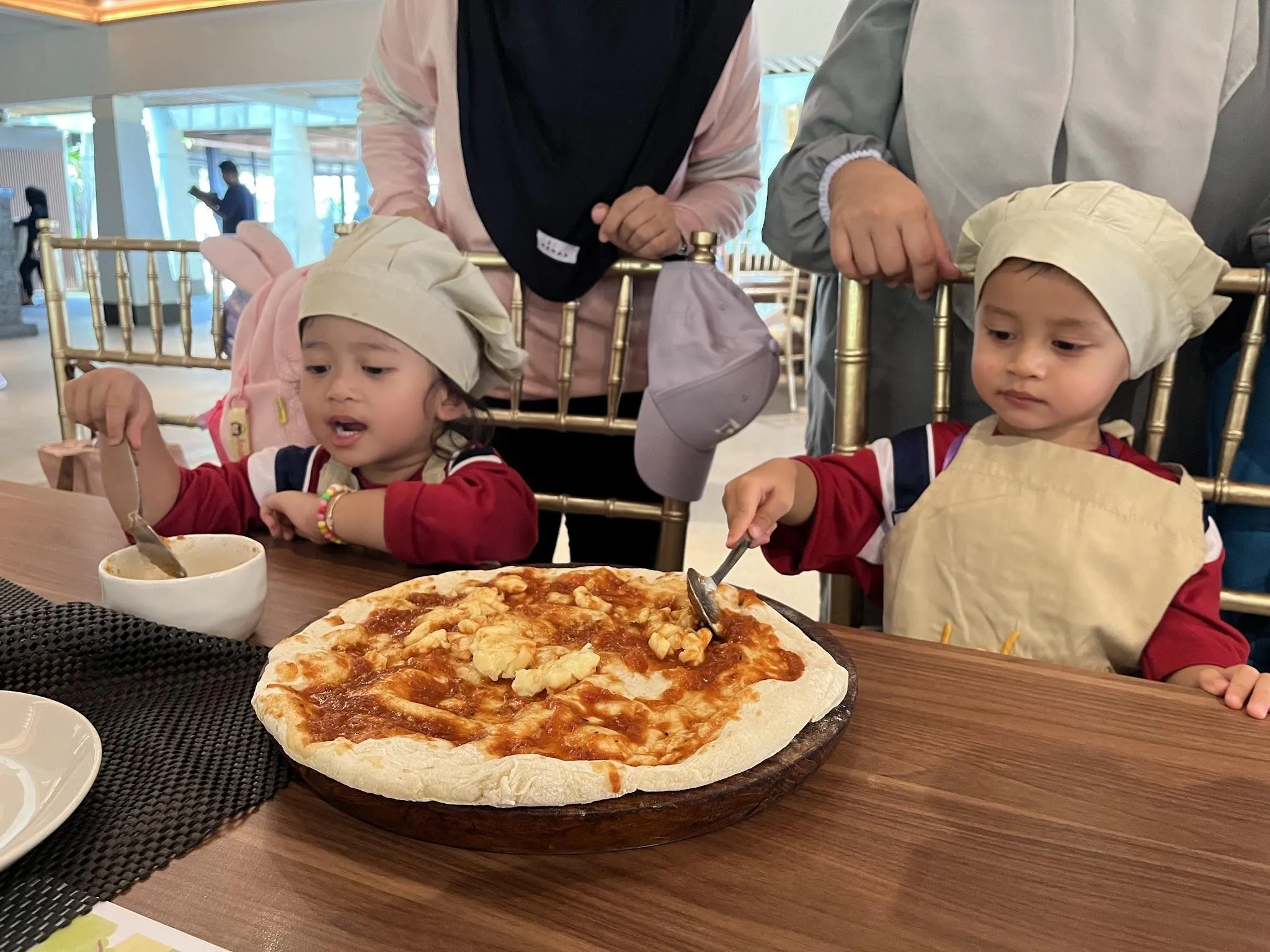 Dua siswa Playgroup bertopi koki mengoleskan saus dan keju pada dasar pizza